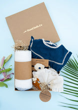 Load image into Gallery viewer, An image of a Baby girl gift hamper with Indigo cotton romper and swaddle, alomg with a hair accessory and a toy, on a light bluish background.