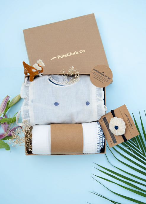 An image of a Baby girl gift hamper with Indigo cotton romper and swaddle, alomg with a hair accessory and a toy, on a light bluish background.