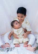 गैलरी व्यूवर में इमेज लोड करें, A boy holding small kid A kid wearing a cotton jumpsuit eco-printed with silver oak leaves on it sitting on a bed.