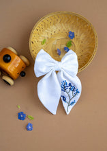 Load image into Gallery viewer, A hair accessory of White Hair Bows - Blue Floral placed in a wooden basket, placed on a light brown background with some tiny blue flowers, leaves and decoratives