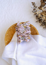 गैलरी व्यूवर में इमेज लोड करें, A hair accessory of Lavender Floral Scrunchies placed in a wooden basket with a white cloth beneath, placed on a white background with some leaves and decoratives