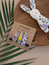 Load image into Gallery viewer, Photo of a set of three Linen Tic Tac Embroidery Hair Clips in pastel colors lying on green leaves on a wooden tray beside a rabbit shaped toy.