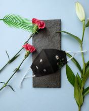 Load image into Gallery viewer, A black color organic cotton hand-embroidered adjustable face masks kept upon a grey sheet with some flowers aside.