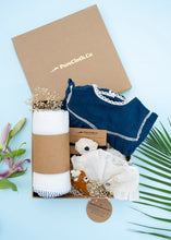 Load image into Gallery viewer, An image of a Baby girl gift hamper with Indigo cotton romper and swaddle, alomg with a hair accessory and a toy, on a light bluish background.

