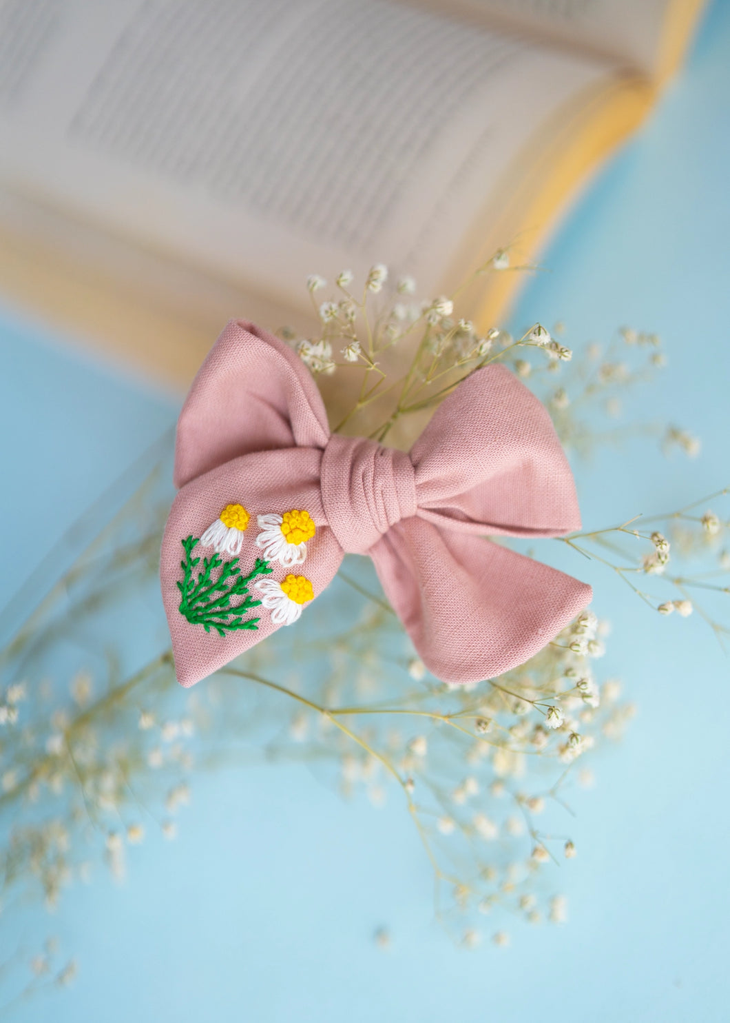 A full size picture of Daisy Delight Knot Hairclip for girls. Hair accessories on cyan background with an open book in the background.