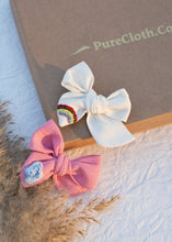 Load image into Gallery viewer, Two Hair Clips with glitters are sitting on a white surface next to flowers and another has paste colored bows with a rainbow
