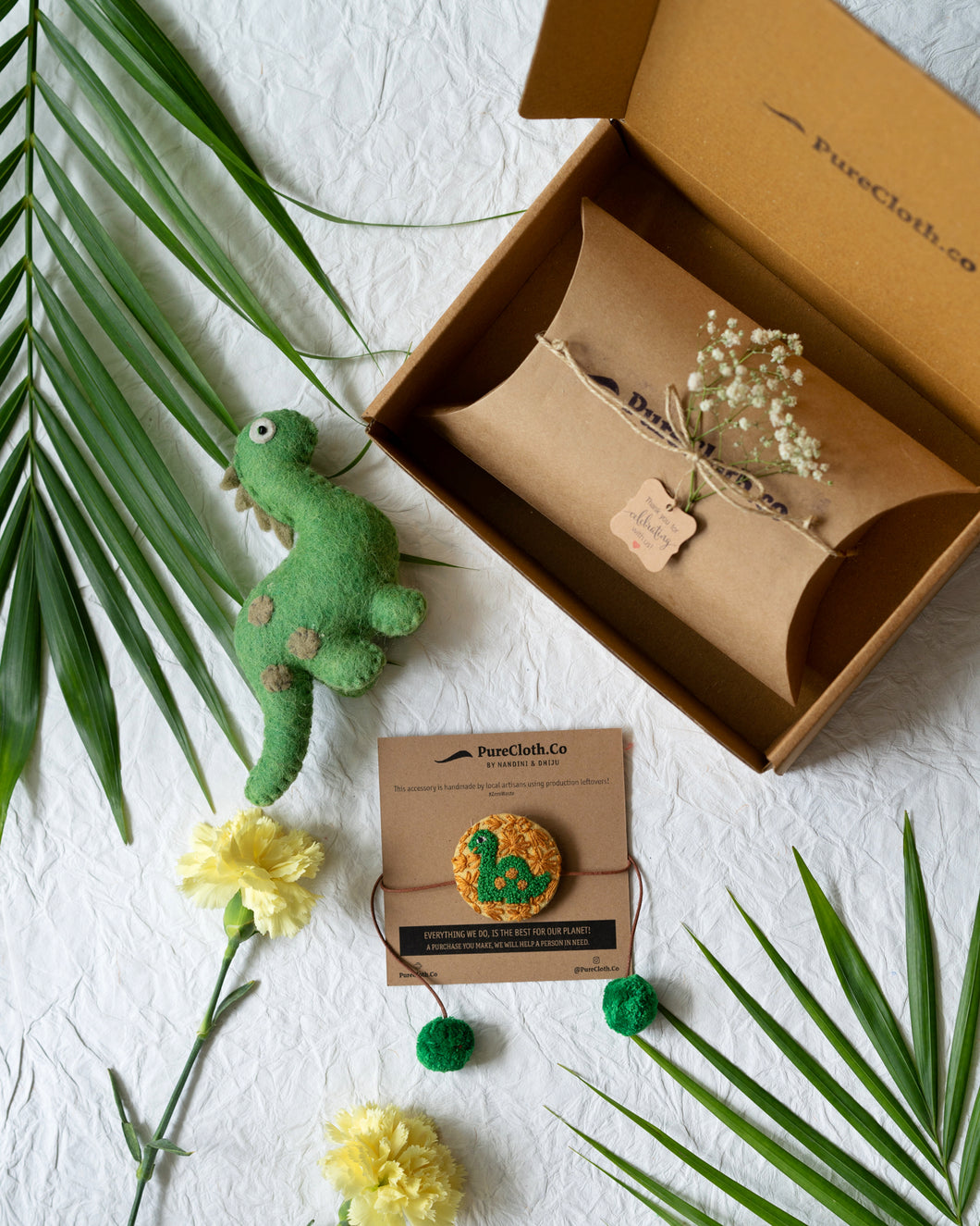 Handmade Dinosaur Rakhi wrapped around brown card with a brown gift box, cute green dinosaur, flower and leaf aside.