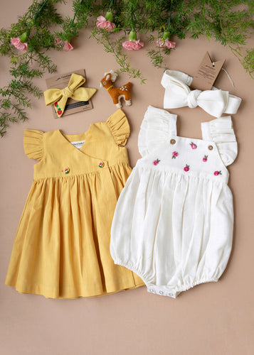 A beautiful pair of organic cotton baby clothes and a  pair of hair accessories  with a cute toy beside it kept on a light peach background with some flower aside.