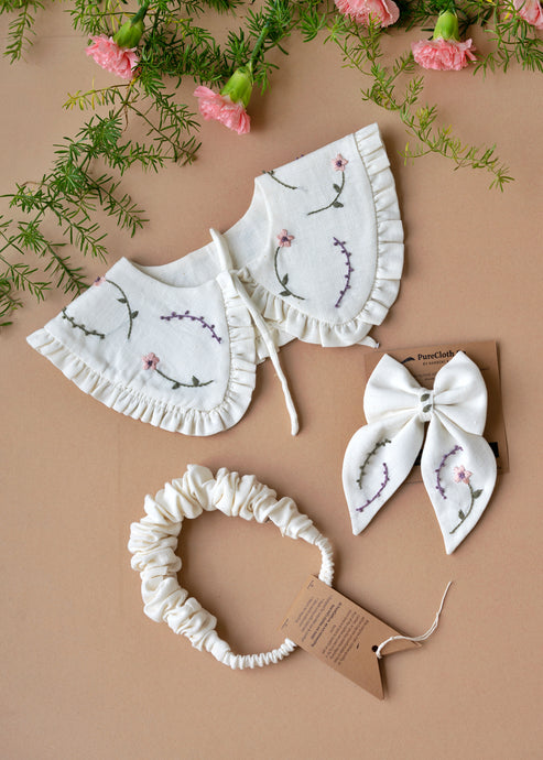 A combo of white and elegant bib, bow and hair head band for kidswear tied upon a brown card kept on peach floor with some flowers aside.