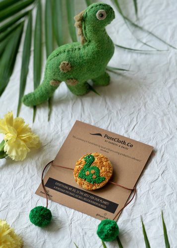 Handmade Dinosaur Rakhi wrapped around brown card with cute green dinosaur, flower and leaf aside. 