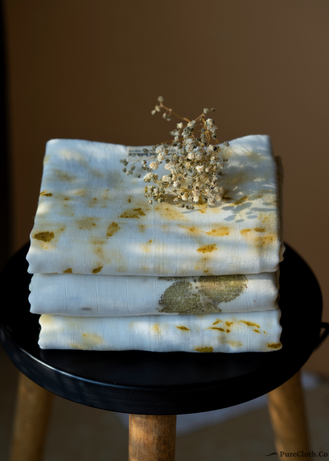 A bundle of muslin baby swaddles with marigold ecoprinted on them kept upon a wooden chair with some flowers upon it.