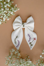 Load image into Gallery viewer, A beautiful white bow with subtle hand embroidery for kids kept on light peach background with some flowers aside.
