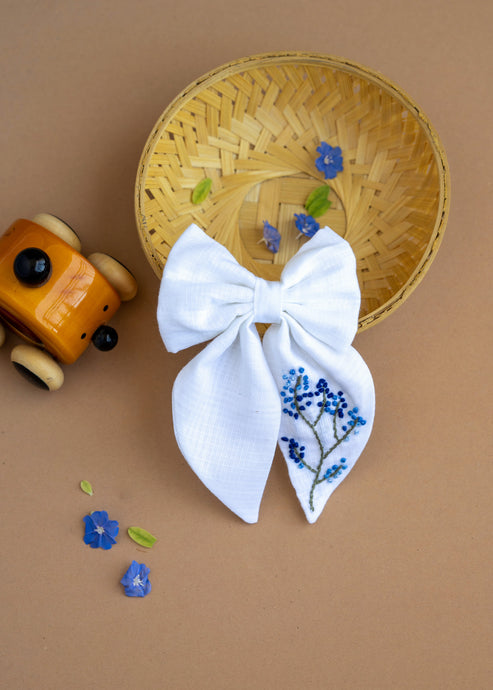 A hair accessory of White Hair Bows - Blue Floral placed in a wooden basket, placed on a light brown background with some tiny blue flowers, leaves and decoratives