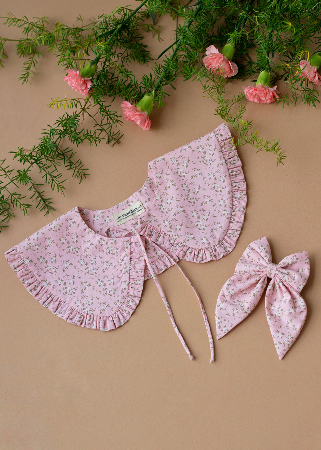 A beautiful pink floral combo of detachable victorian style collar and matching bow with small frills around it for kids kept on light peach background with some flowers aside.