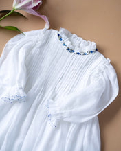 Load image into Gallery viewer, The pintuck detailing across the bust and ruffles running across the neckline in classic white palette kept upon a peach background with a leaf aside.
