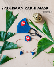 Load image into Gallery viewer, A blue handmade organic cotton spiderman rakhi with mask kept on a peach background with some leaves aside.
