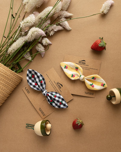 A beautiful designer hair accessories combo tied upon a brown card with some strawberry, toy bell and flowers around it.