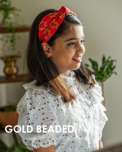 Load image into Gallery viewer, A young and beautiful girl wearing gold-beads hand crafted on red cotton fabric hair accessories showing her side profile and with some plants stand in the background.
