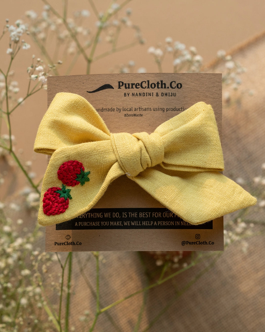 A yellow knotted ribbon bow hair clip with strawberry design on it wrap on a brown card with some flowers in background.