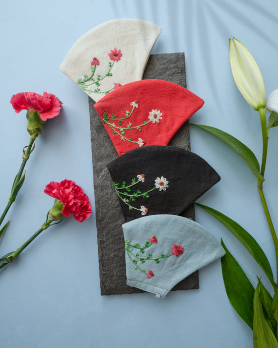 A different color organic cotton hand-embroidered adjustable face masks kept upon a grey sheet with some flowers aside.