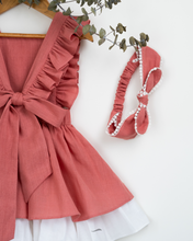 Load image into Gallery viewer, A back view of a beautiful dress with a criss-cross back hung on a hanger with a matching hair accessory beside it with a stem of leaves upon it.
