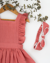 Load image into Gallery viewer, A beautiful dress with a criss-cross back hung on a hanger with a matching hair accessory beside it with a stem of leaves upon it.
