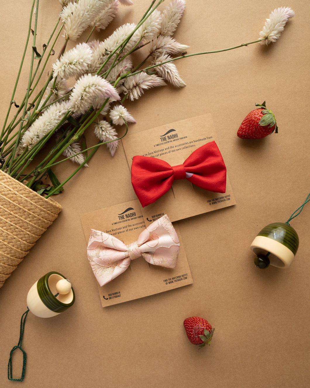 A red and baby pink silk hair clip combo attached on a brown card with some toy bells and strawberries and a bunch of flowers kept aside.