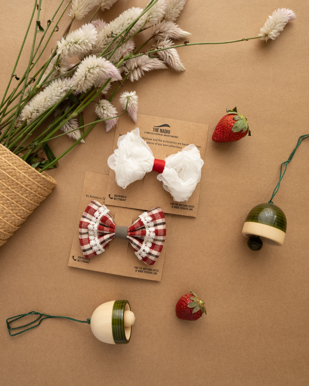 Beautiful white and red hair clip attached to brown card with strawberries, bell toys and bunch of flowers aside.